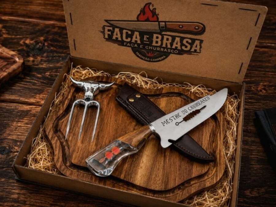 Faca de churrasco e tábua de madeira dentro de caixa artesanal, acompanhadas de garfo e bainha de couro.